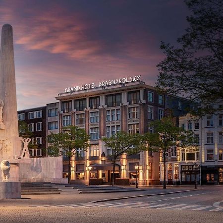 Anantara Grand Hotel Krasnapolsky Amsterdam Buitenkant foto The photo depicts a city square scene featuring a monument or sculpture in the foreground, made of light-colored stone. The monument is tall and appears to feature figures or carvings. Behind the monument, there is a large hotel building, identified 