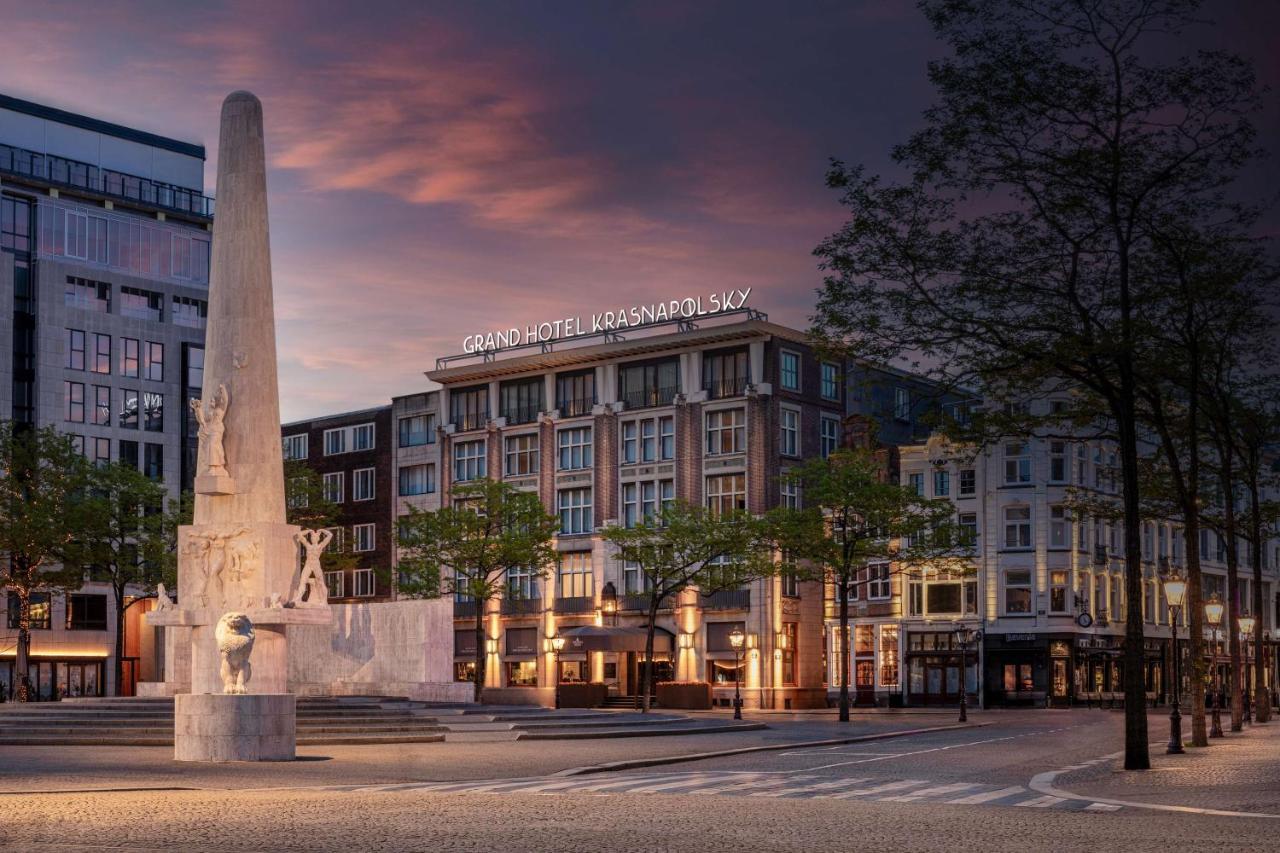 Anantara Grand Hotel Krasnapolsky Amsterdam Buitenkant foto The photo depicts a city square scene featuring a monument or sculpture in the foreground, made of light-colored stone. The monument is tall and appears to feature figures or carvings. Behind the monument, there is a large hotel building, identified 