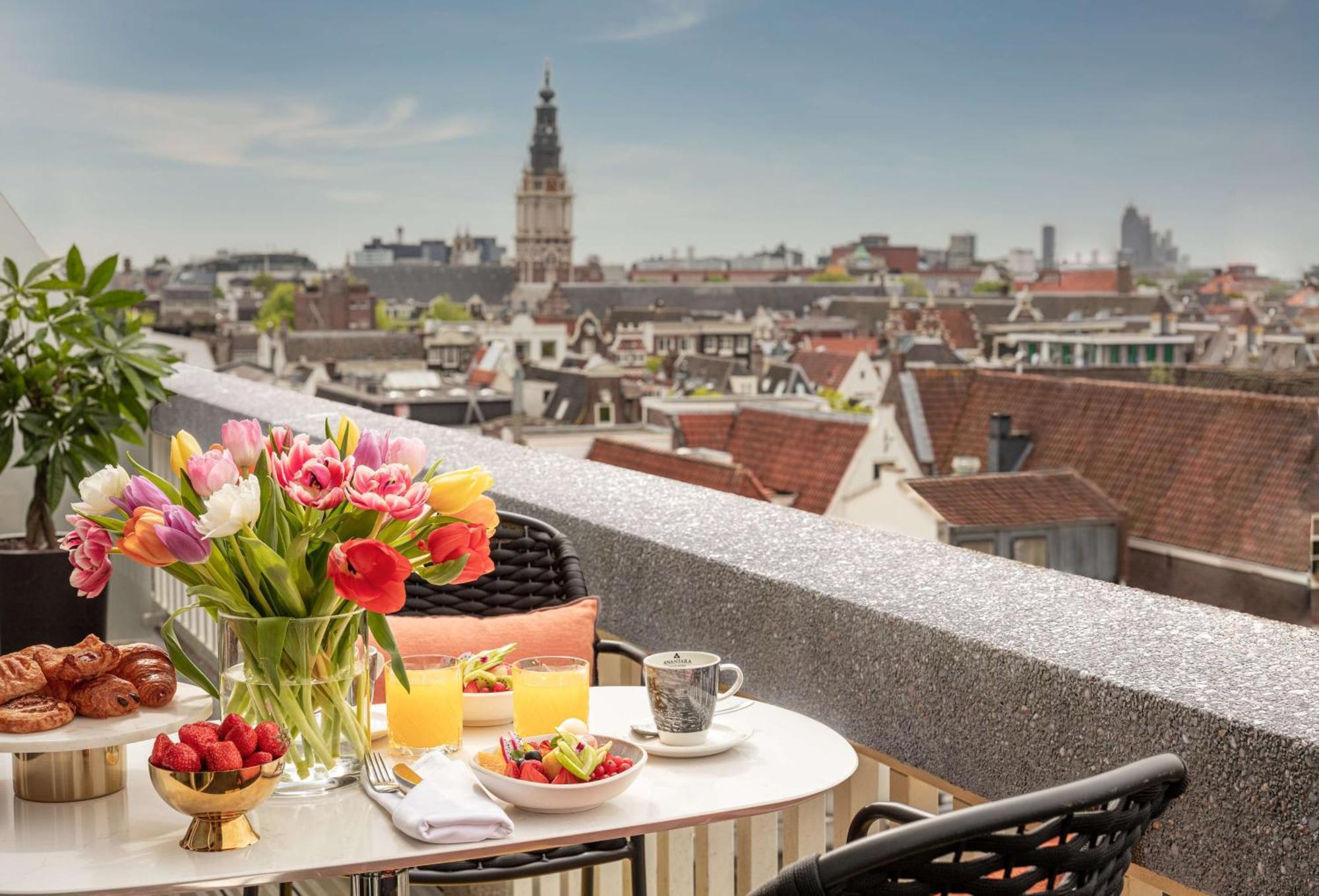 Anantara Grand Hotel Krasnapolsky Amsterdam Buitenkant foto The photo displays a beautiful balcony setup overlooking a cityscape. A round table is set for breakfast, featuring a bouquet of colorful tulips, a plate of fresh fruits, pastries, and two glasses of orange juice. There’s also a cup, possibly for cof